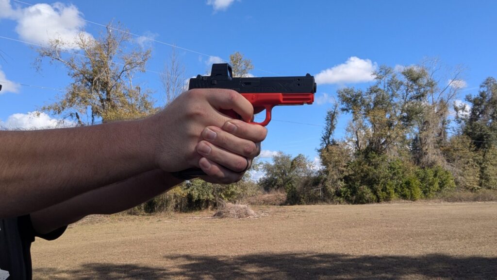 A side profile of a man's hands holding a red Mantis training pistol in a standard shooting stance against a field background.