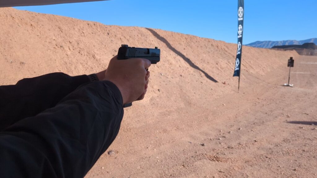A person holding a black handgun at a range, seen from behind their hands under a branded tent.