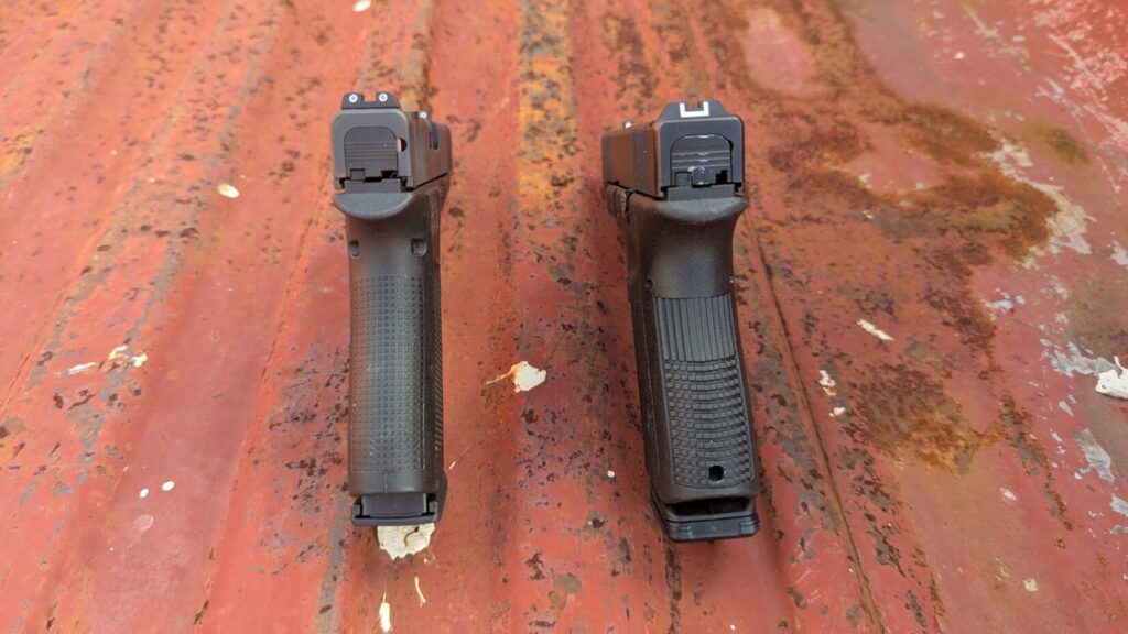 Rear view of two black semi-automatic pistols showing different grip widths and textures on a rusty red surface.