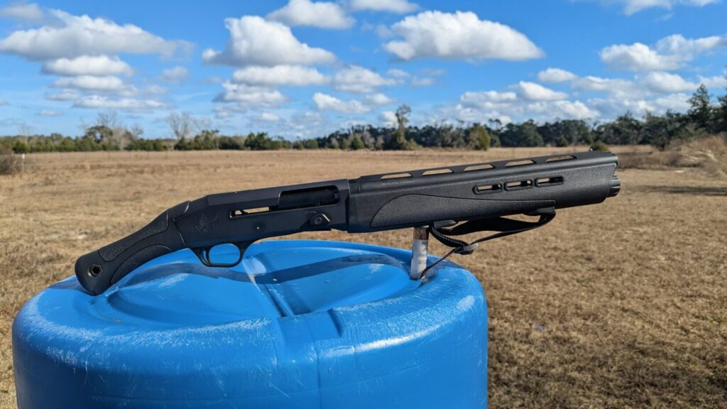 The IWI Mafteah balanced on a blue plastic barrel with a wide-open field and cloudy sky in the background.