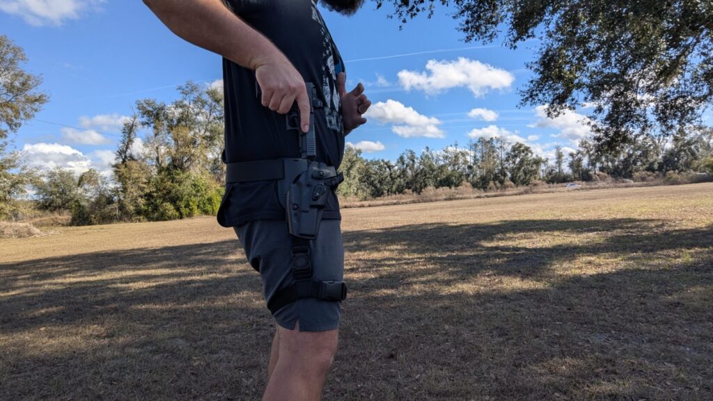 A person standing in an open field reaching down to draw a Glock 17 Gen 6 from a black mid-ride duty holster attached to a belt.