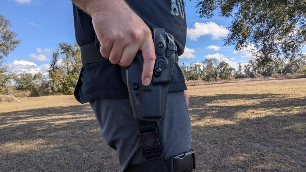 A person standing in an open field reaching down to draw a Glock 17 Gen 6 from a black mid-ride duty holster attached to a belt.