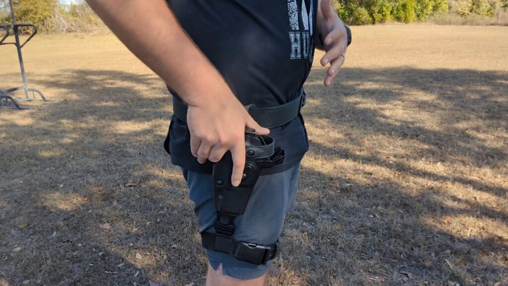 A side-view close-up of a hand resting on a handgun that is secured in a black tactical holster attached to a leg rig.