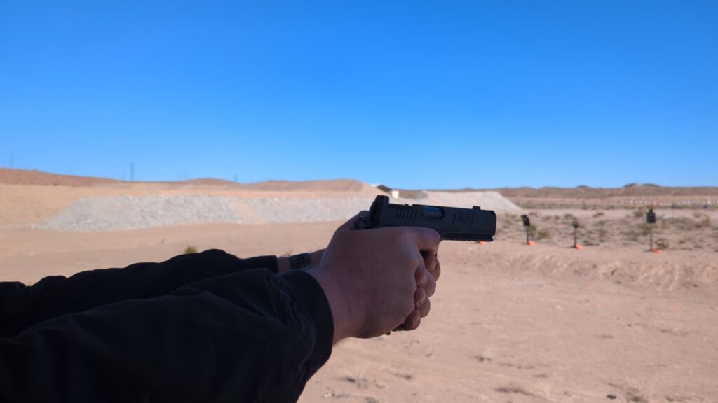 Point-of-view shot of a person aiming a black handgun at targets in an outdoor desert shooting range.