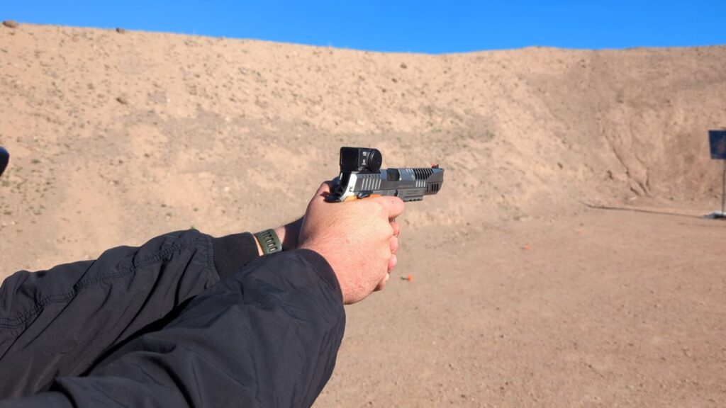 POV of a person aiming a silver handgun equipped with a red dot sight at an outdoor desert range.