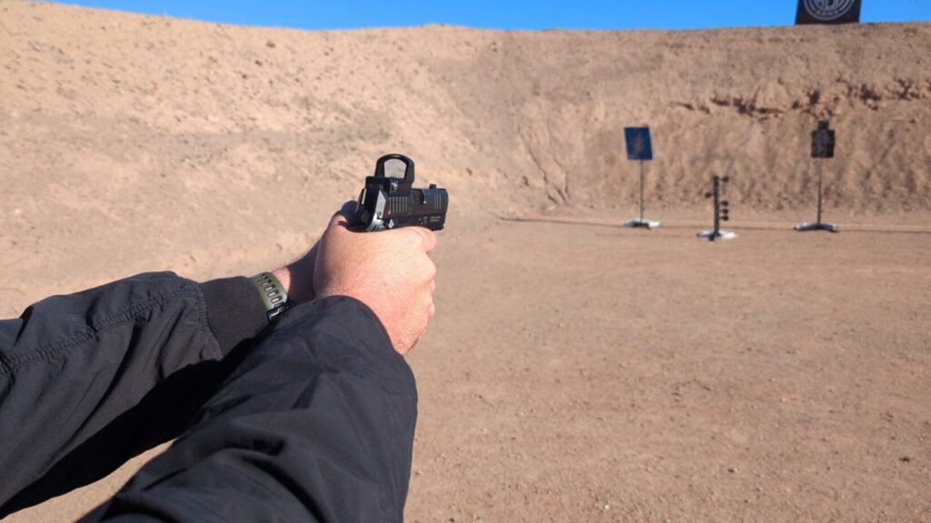 Point-of-view shot aiming a black handgun with an optic toward steel targets in a desert environment.