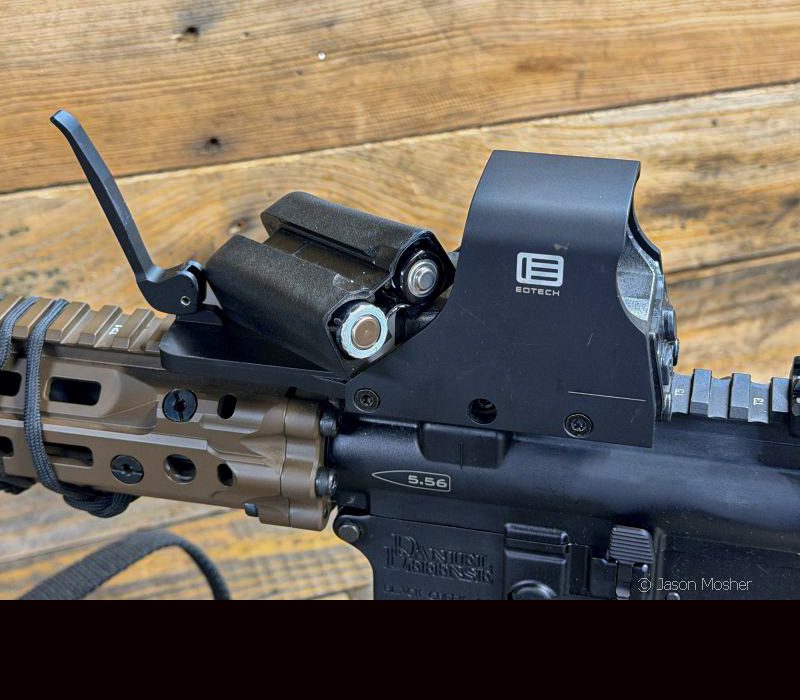 A black optic mounted on a rifle with the battery compartment pulled out exposing the batteries inside it. 