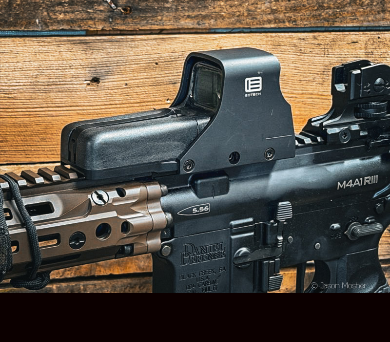 A view of the side of a black Eotech optic mounted to a rifle with a wood wall in the background. 
