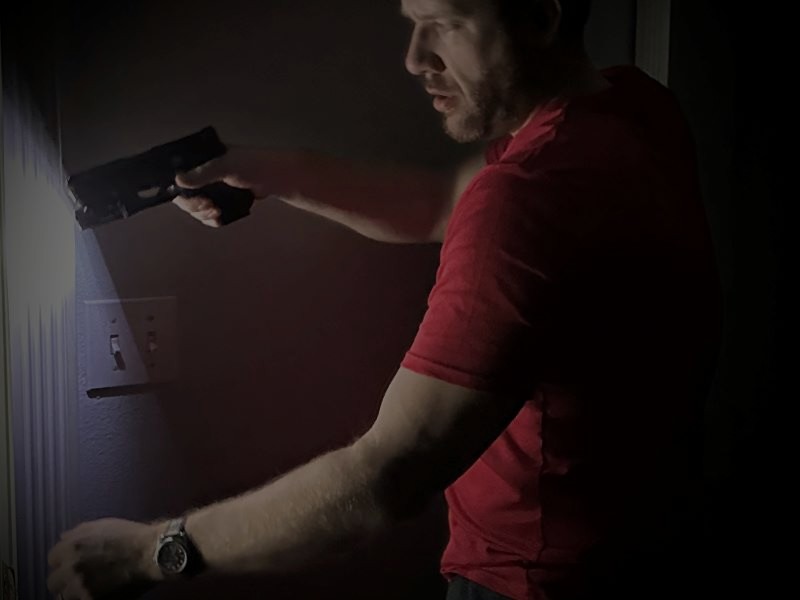 A person in a red shirt looking through a door of a dark room, using a black handgun and alight mounted to the handgun.