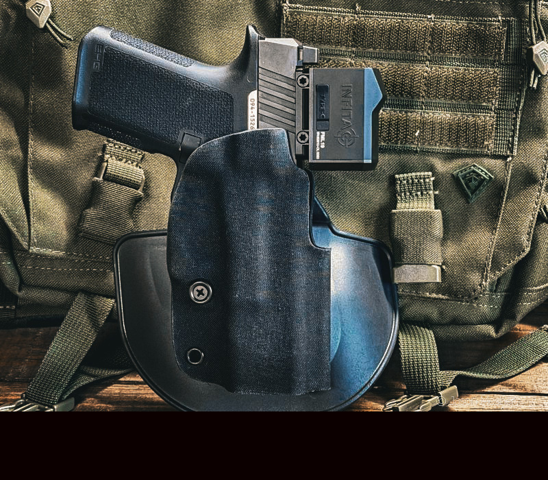 A black RXM pistol with a black thermal optic mounted to it, in a black holster in front of a green vest. 