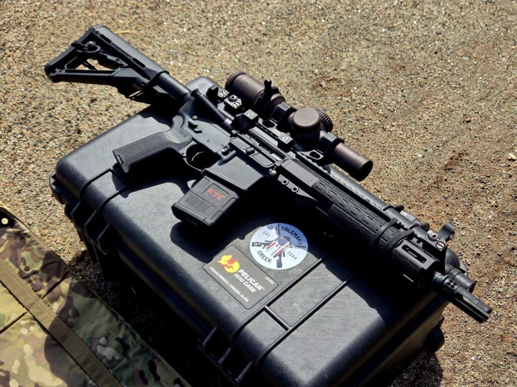 A rifle with a LPVO optic on top of a pelican case against a dirt background