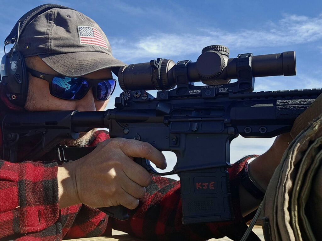 A man in a red flannel shirt and American flag ball cap takes aim with a rifle on a rest