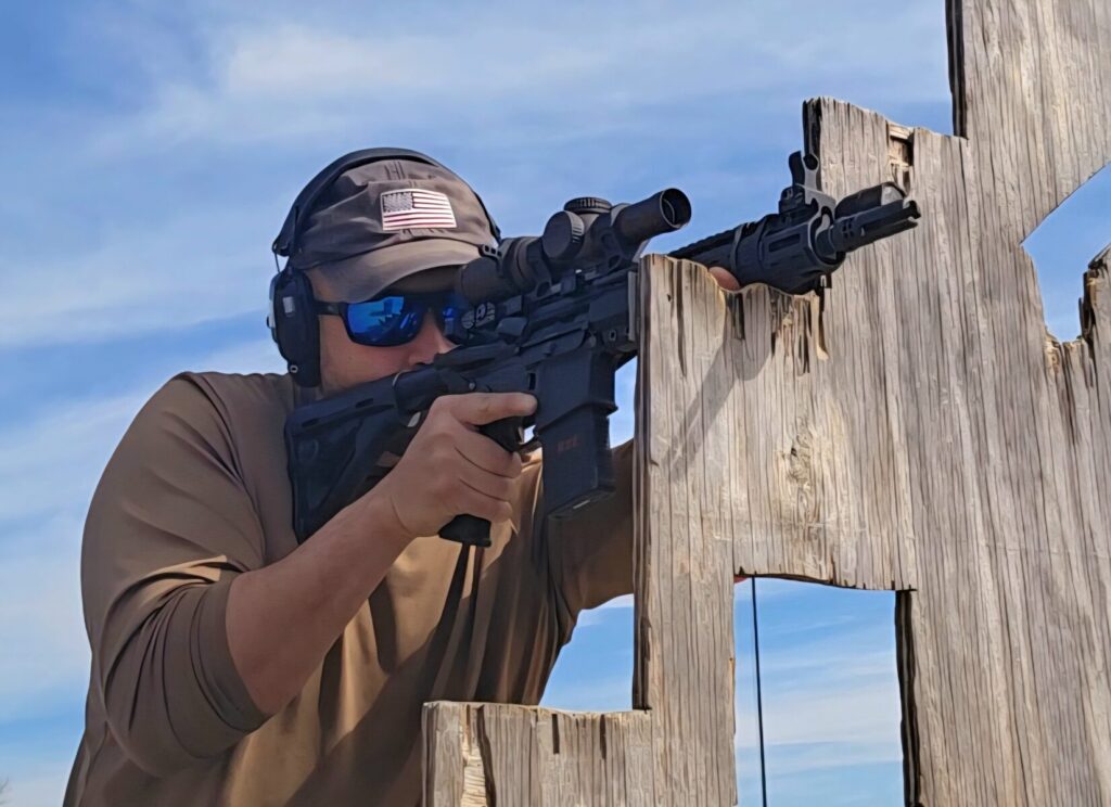 A man in an American flag ball cap holds a rifle on a VTAC barricade