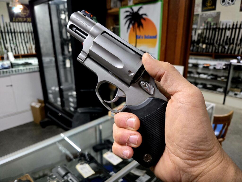 A large revolver in a man's hand with firearms in the background on racks