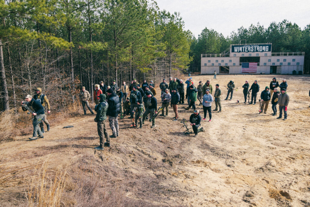 A group of bowhunters practice in a field with a connex container superstructure in the background