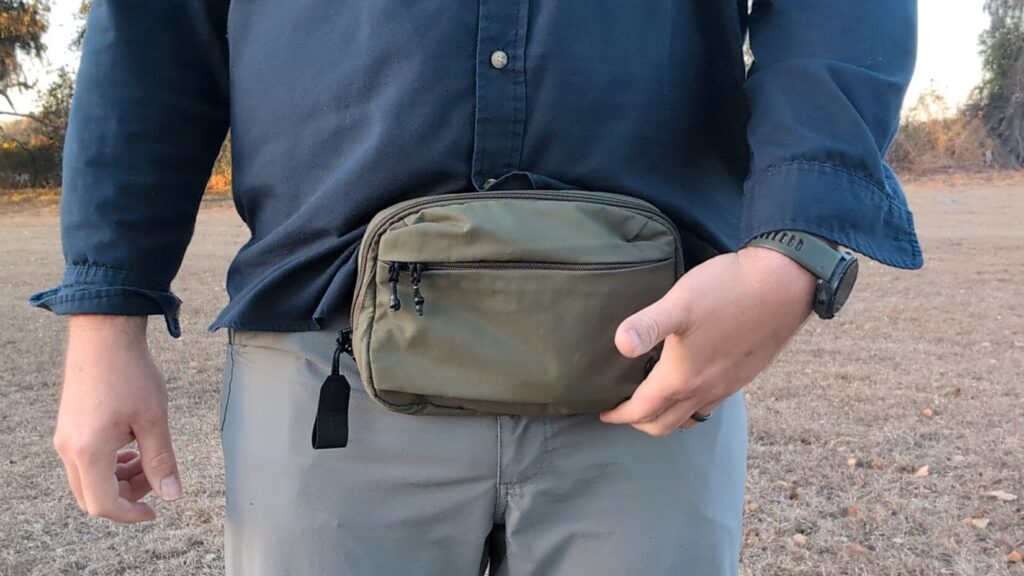A person rests their hand on the bottom of the olive green waist bag, showing its position relative to the waistband.