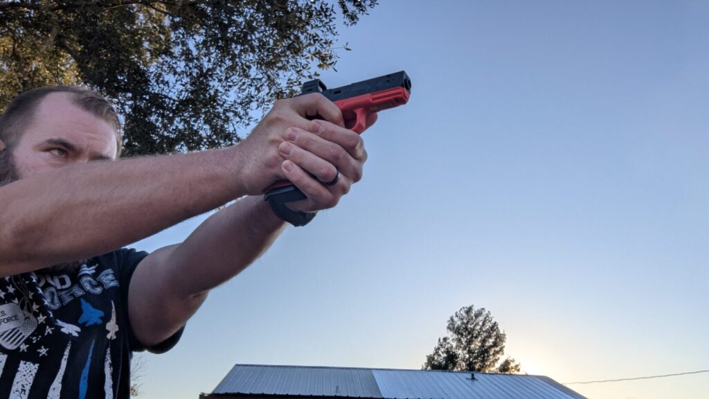A shooter holding the red Mantis training pistol in a two-handed grip, aiming upward against a clear blue sky