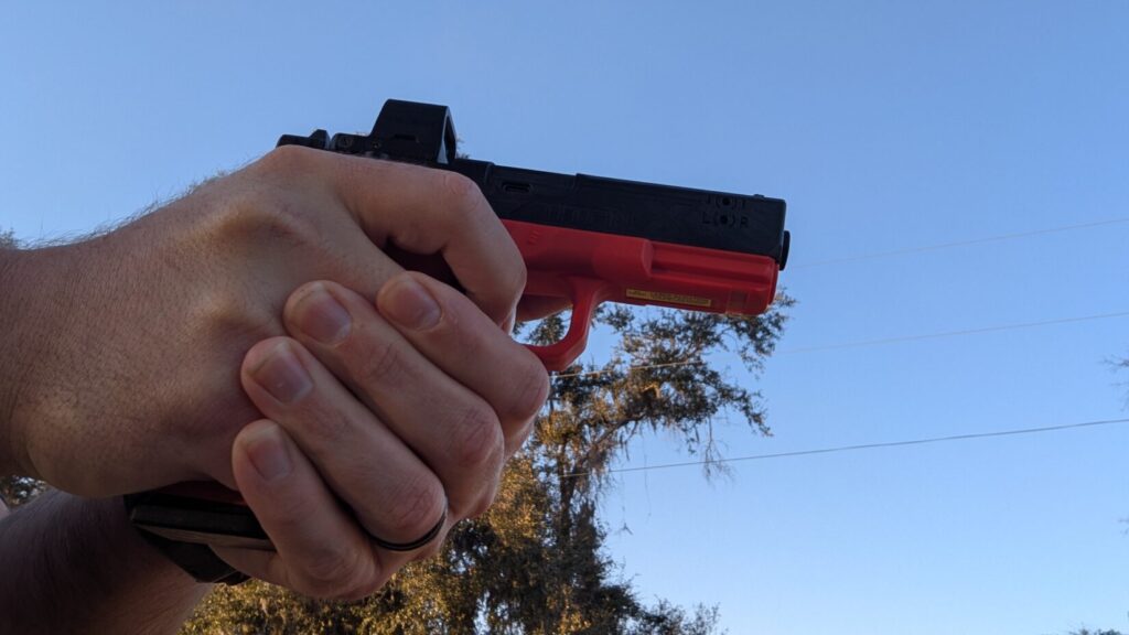 person's hands holding a black and red training handgun with an optic, with their right thumb extended forward along the frame.
