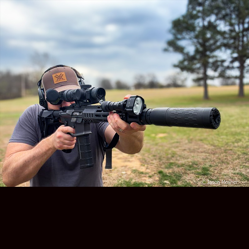 A person in a grey shirt and hat, shooting the MK1 rifle with a silencer attached. 