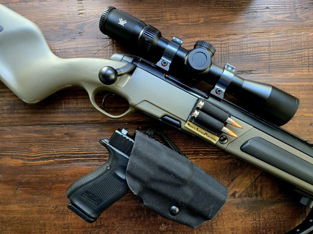 A bolt action rifle with optic and spare ammunition next to a handgun in a black holster on a wooden backdrop