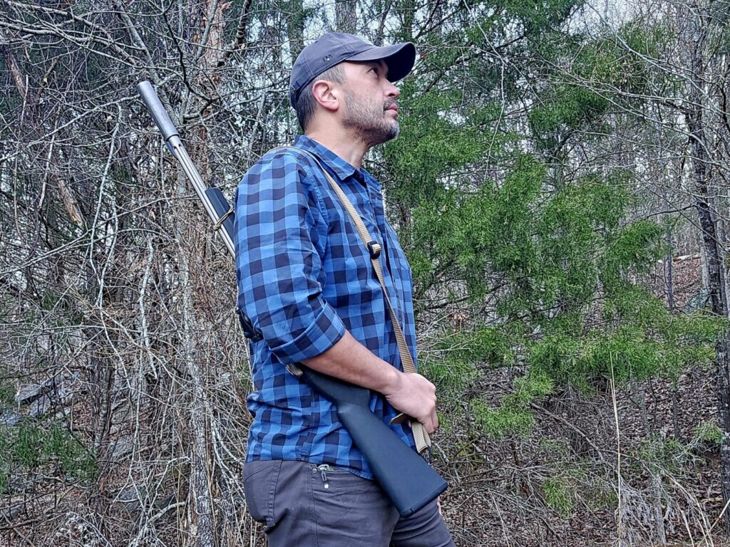 A man in a blue plaid shirt with a slung rifle over his shoulder