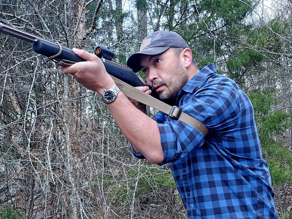 A man takes aim with a rifle in a wooded setting