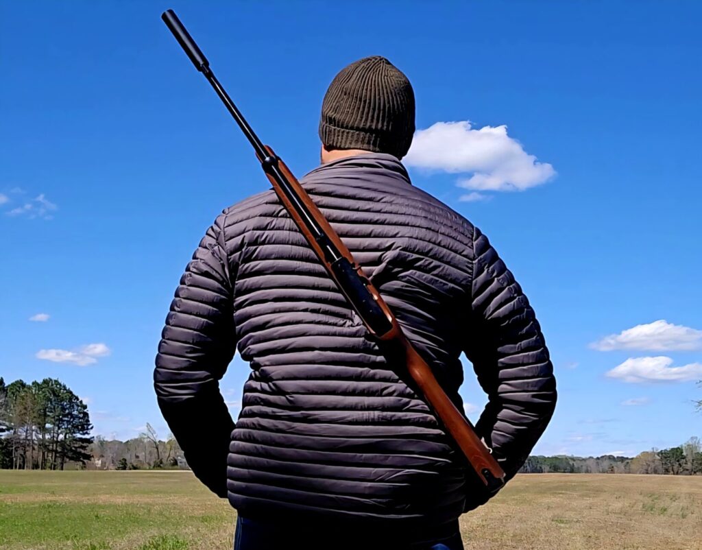 A man in a gray jacket with a green hat stands at the edge of a field with a rifle across his back.