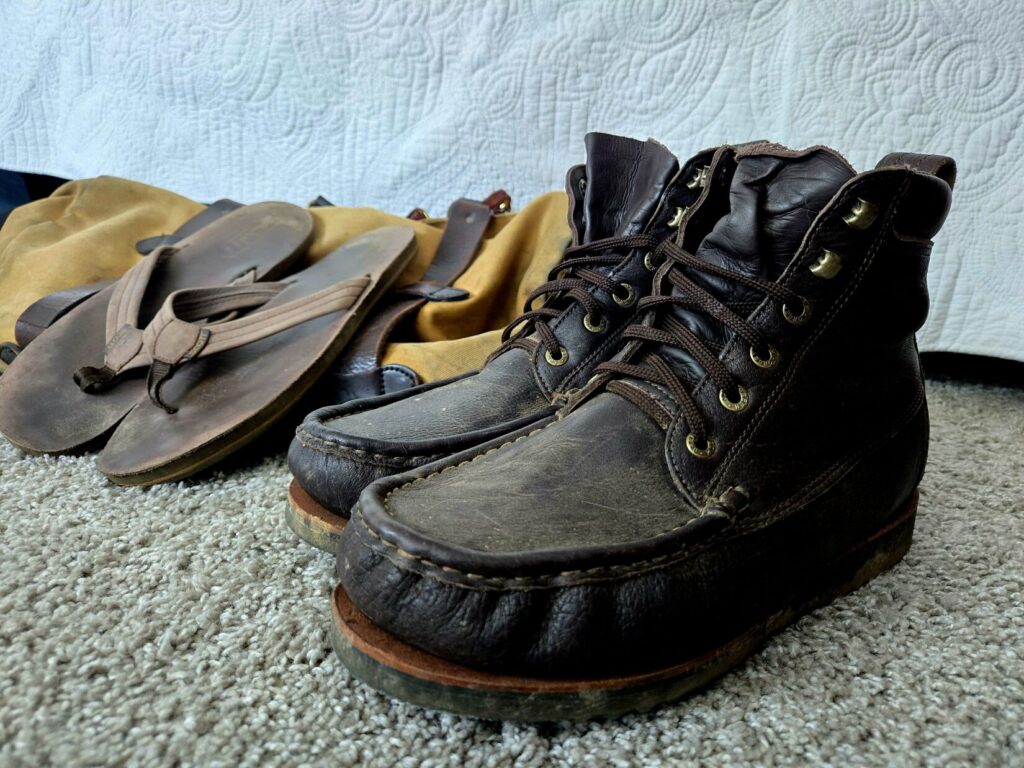 A pair of boots and sandals on top of carpeting next to a bed with a bag in the background.