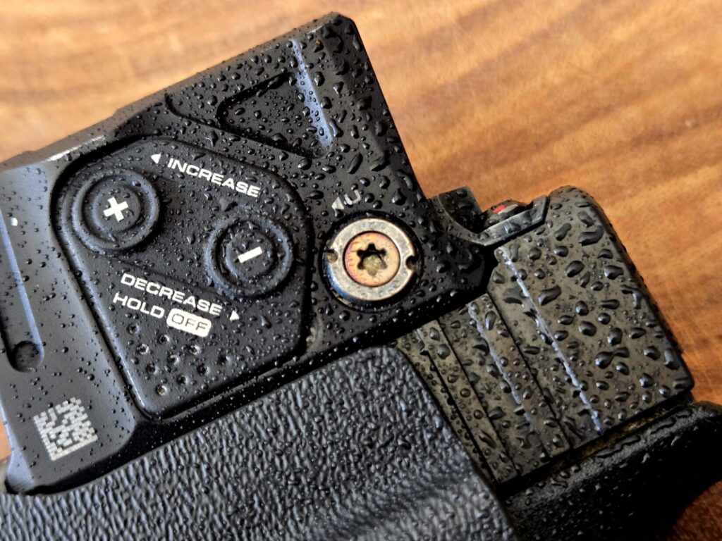 Beads of moisture on a pistol optic against a wood background