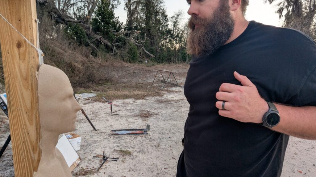 A close-up side view of a shooter standing in front of a training mannequin, with his non-firing hand indexed against his chest to maintain a clear path for a close retention draw.