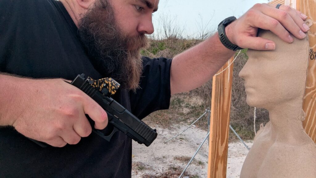 A shooter moves through a sandy range while maintaining a high-ready retention position, keeping the handgun close to the body for weapon retention.
