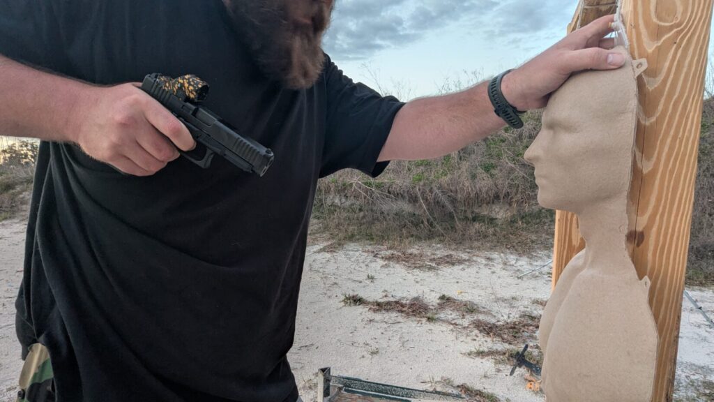 A shooter practices a close retention position, holding a handgun tight against their ribs with one hand while the other hand creates distance by pushing against the head of a training mannequin.