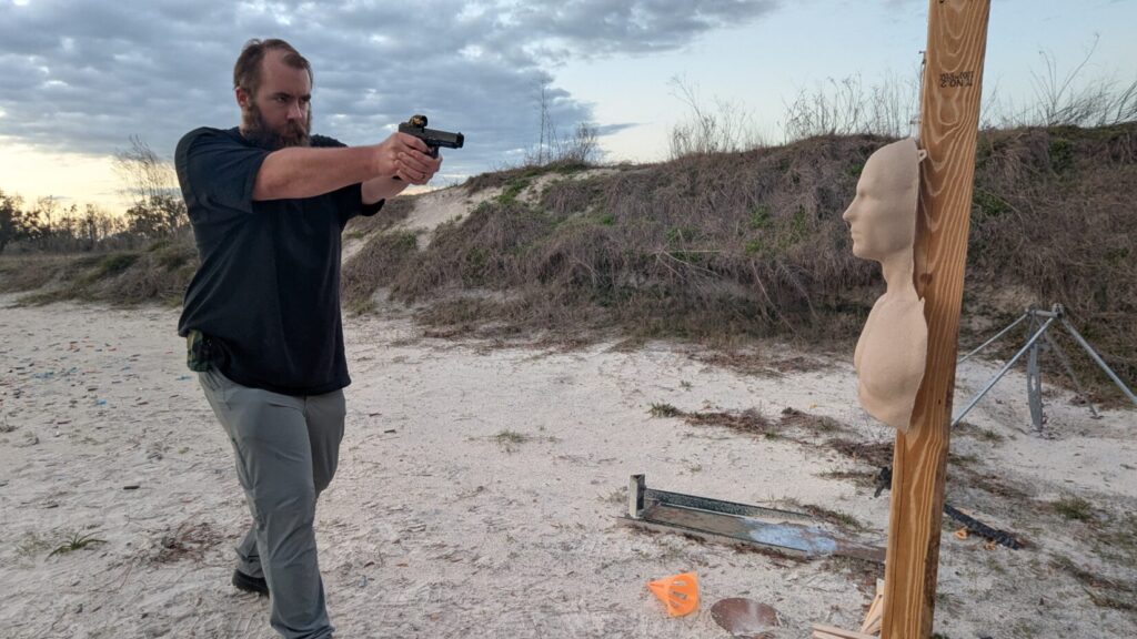 A shooter stands at a sandy range, extending a handgun with both hands to aim at a training mannequin mounted on a wooden post.