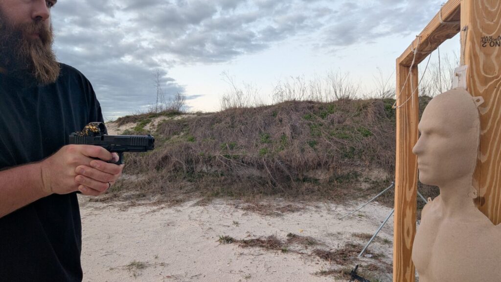 A shooter holds a handgun in a compressed ready position at chest level, facing a training mannequin in a sandy outdoor range environment.