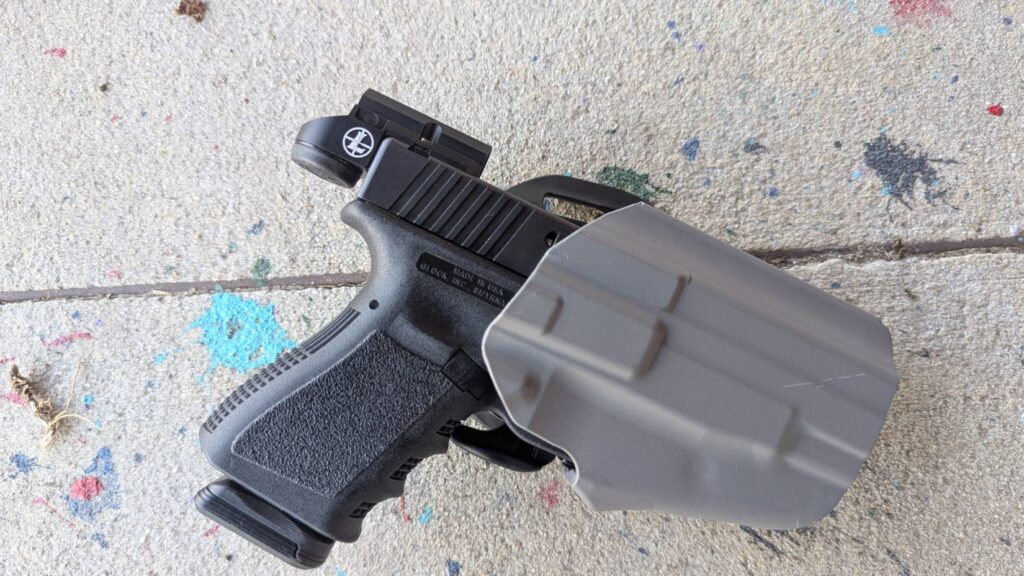 A Glock with a Leupold DeltaPoint Micro optic shown inside a grey Safariland holster, resting on a concrete floor with paint splatters.