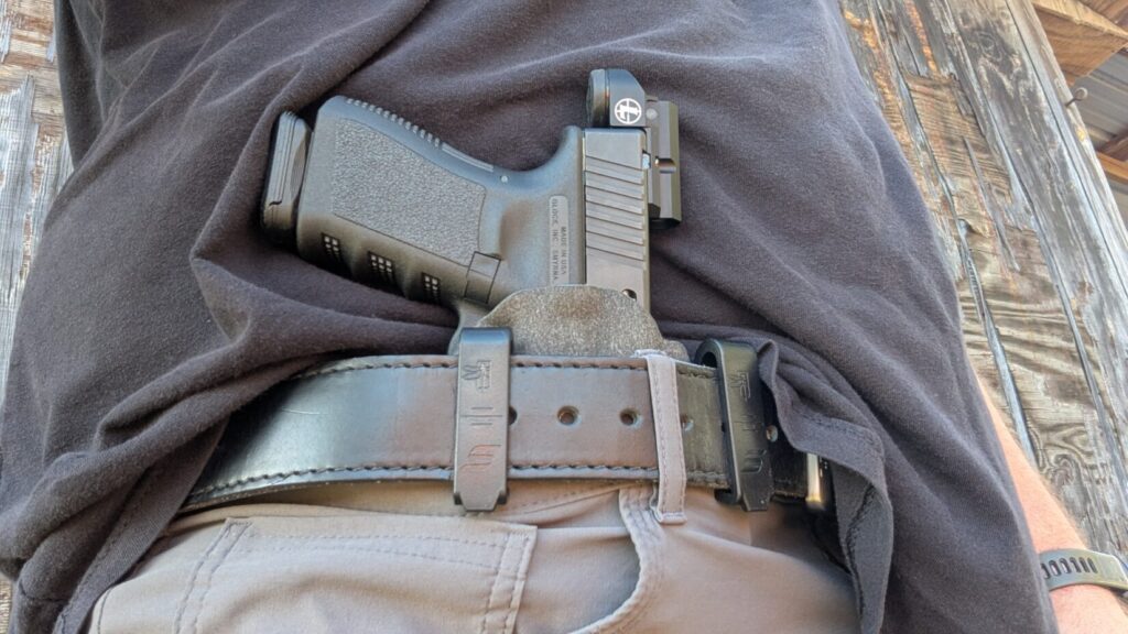 A Glock equipped with a Leupold DeltaPoint Micro carried in an appendix IWB holster, tucked into a waistband with a leather belt.