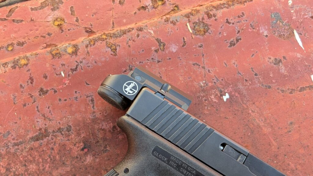 Profile view of a Glock handgun resting on a rusted red metal surface, featuring the low-profile Leupold DeltaPoint Micro mounted to the rear of the slide.