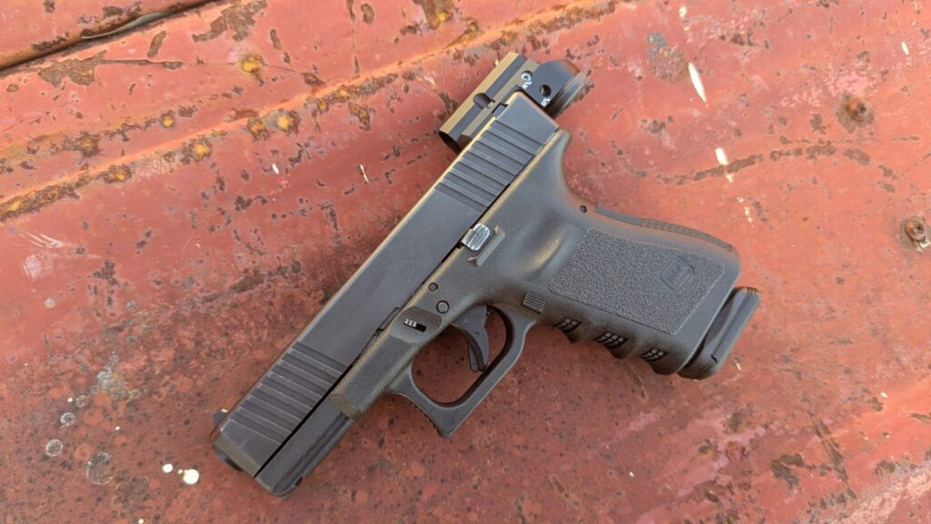 A full side view of a Glock pistol equipped with the Leupold DeltaPoint Micro, resting on a wooden surface outdoors with a field and trees in the background.
