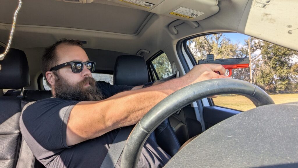 A man in the driver's seat aiming a red training pistol toward the side window