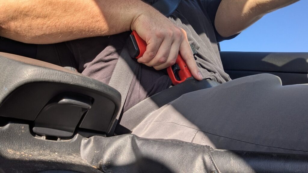 A red training pistol with an optic sight shown concealed in the waistband, partially covered by a shirt and seatbelt.