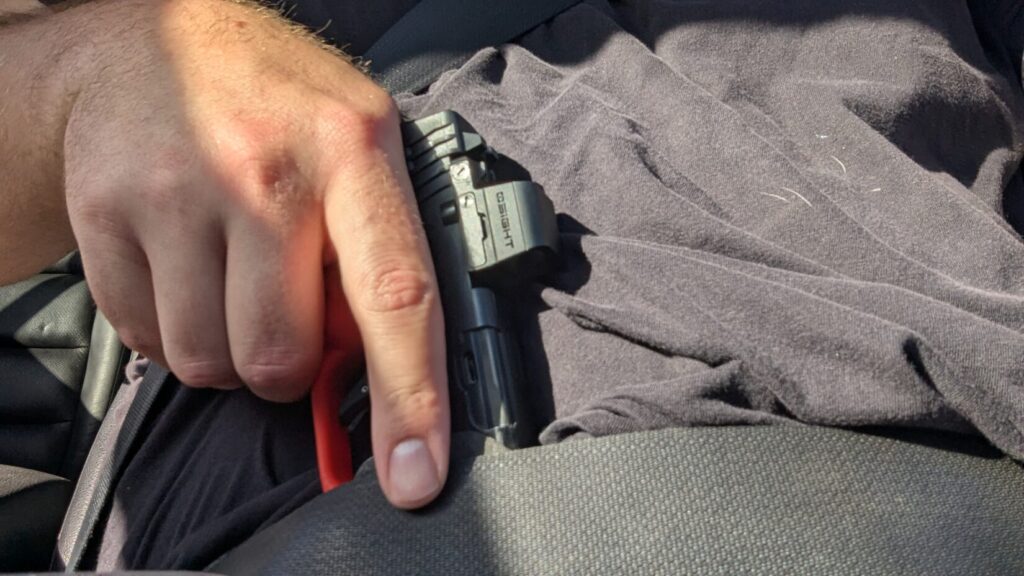 Close-up of a person gripping the handle of a red training pistol tucked under a seatbelt for a draw.