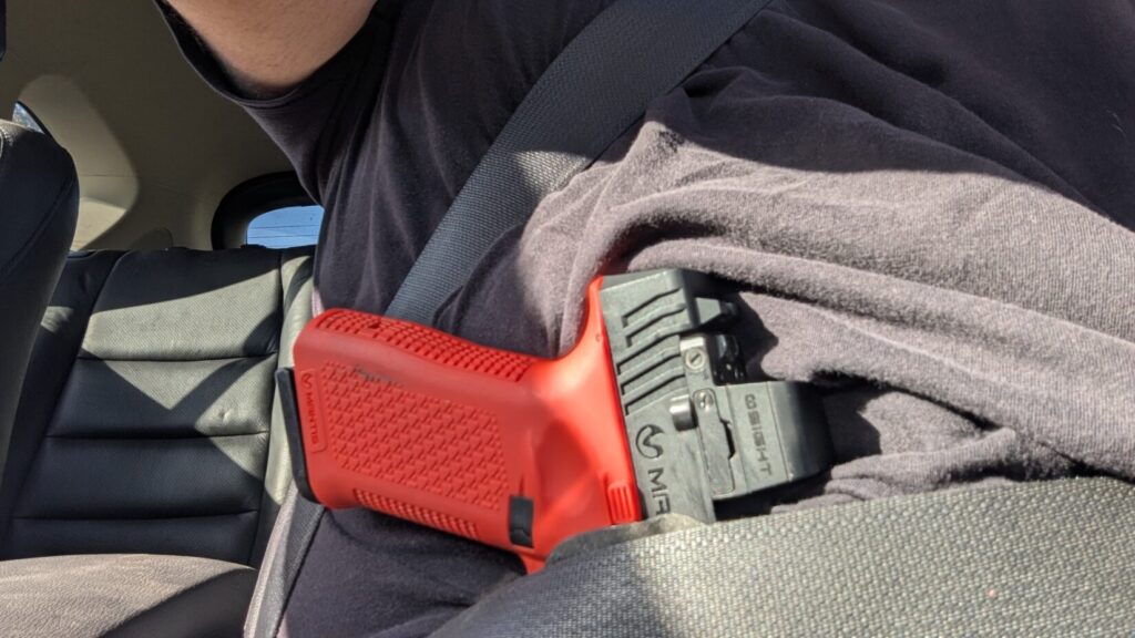 Close-up of a hand clearing a t-shirt to access a concealed training pistol tucked into the waistband behind a seatbelt.