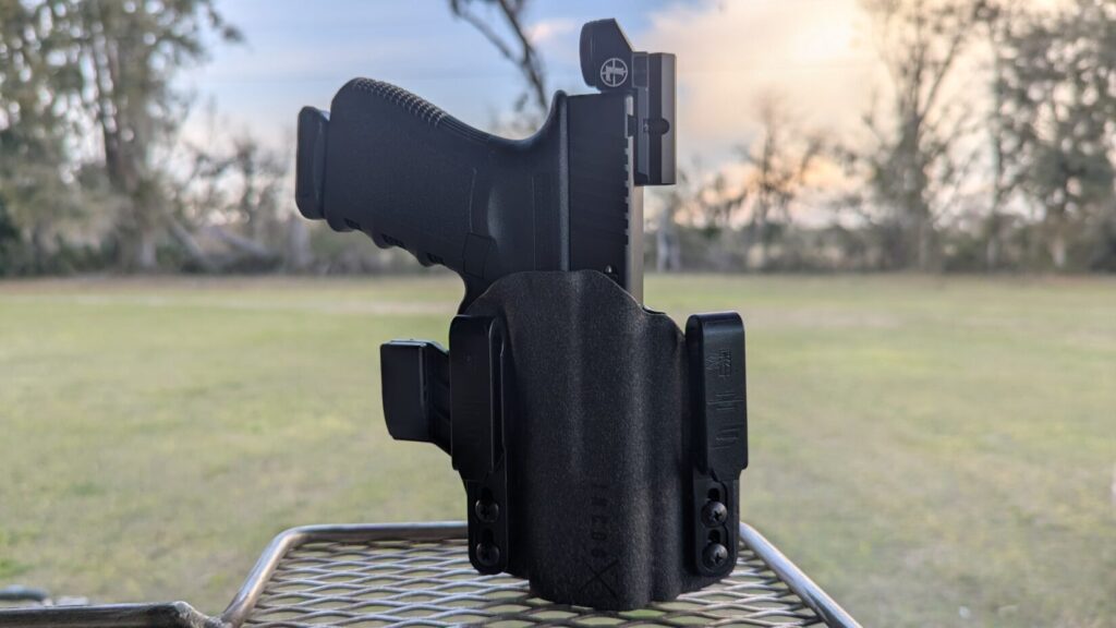 A Glock handgun with a red dot sight secured in a black Kydex IWB holster, standing upright on a metal mesh table.