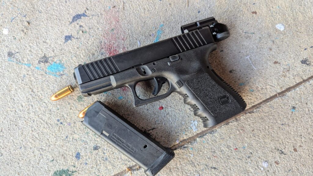A Glock with an optic mounted, leaning against a wall next to a loaded magazine and a single standing bullet.