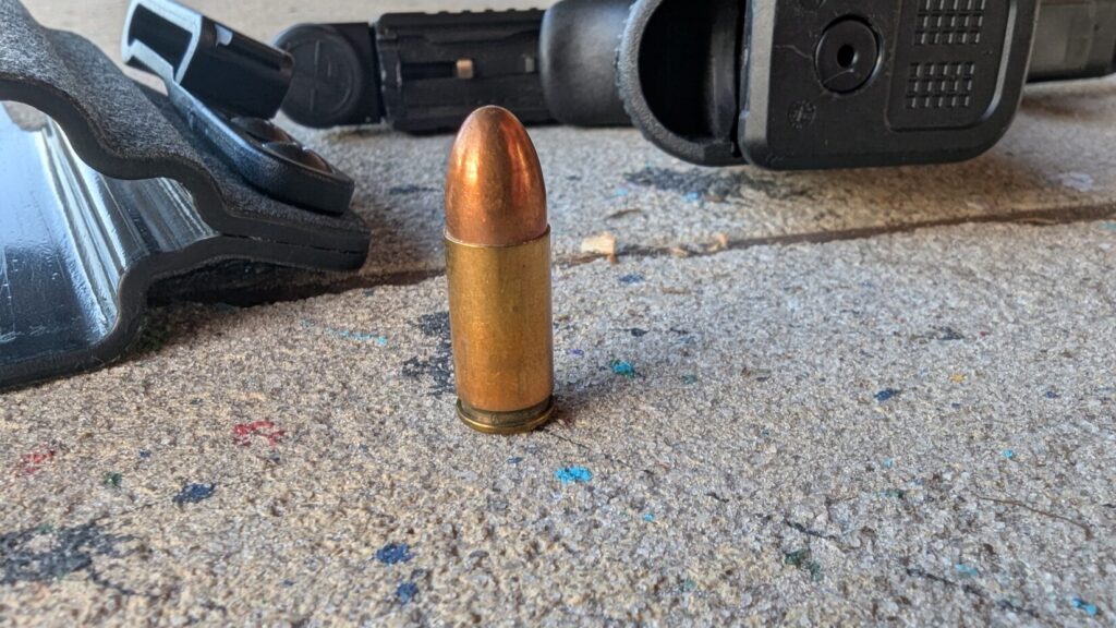 A single 9mm round standing upright on a concrete surface next to a handgun and holster.