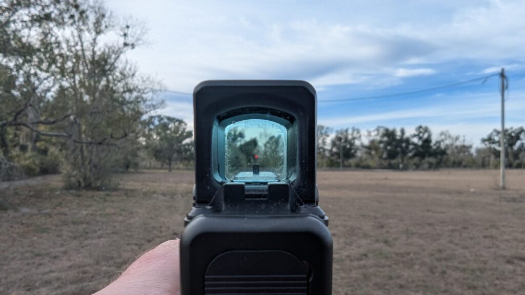 First-person view through the Aimpoint COA window, showing the bright red aiming dot centered in an open field.