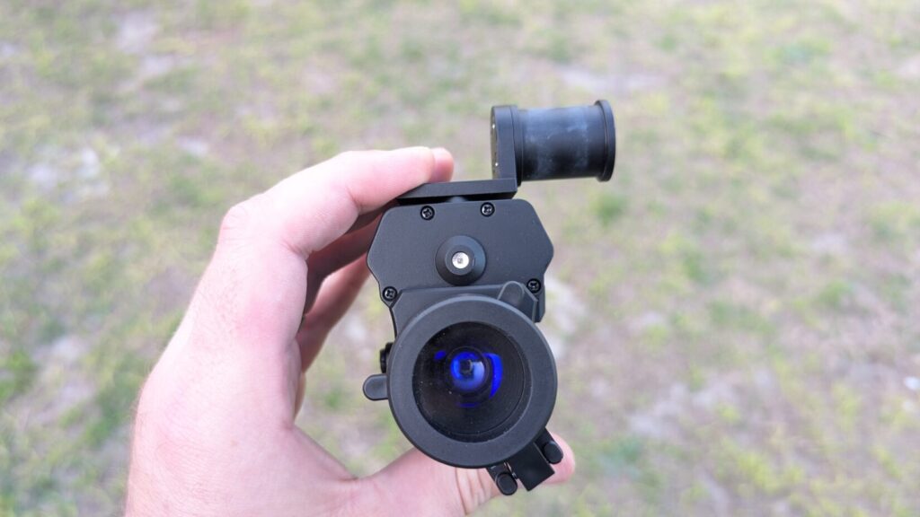 Front view of the Goodnite Gear NVG 50 held in hand, showing the objective lens with a blue-coated tint and a top-mounted dovetail adapter.