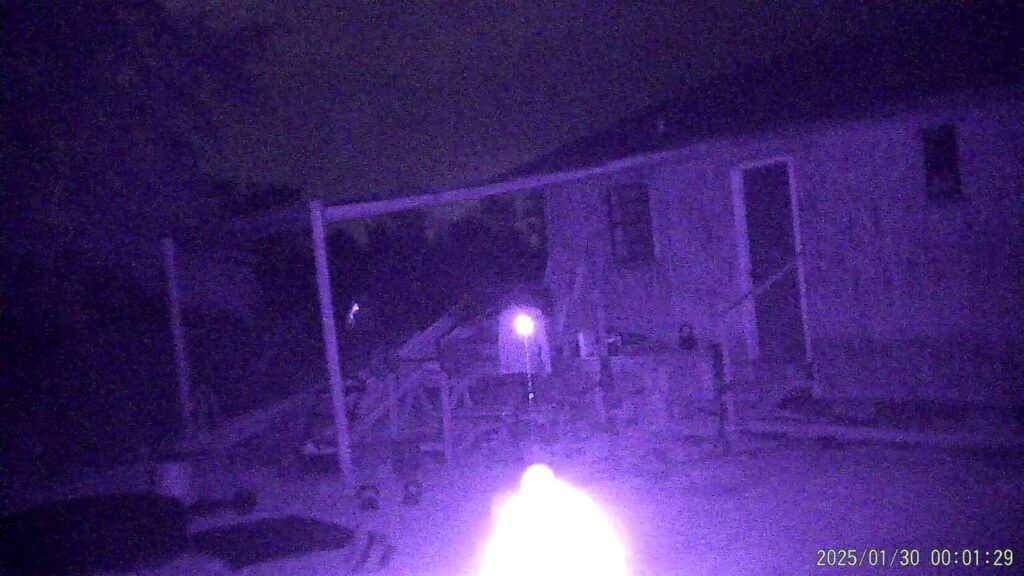 A purple-tinted night-vision view of a backyard structure or porch area. A bright, blooming light source in the foreground creates a heavy glare, partially obscuring a building and wooden framing in the background.