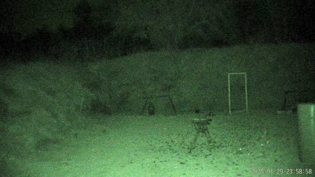  A grainy, green night-vision shot of an outdoor range or field. A white rectangular target frame stands on the right, with a small tripod or stool positioned in the center of the clearing.