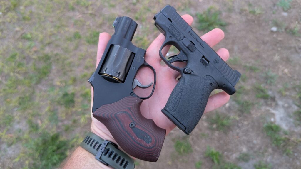 A hand holding a Smith & Wesson snub-nosed revolver with wood-patterned grips above a Smith & Wesson Bodyguard 2.0 semi-automatic pistol for a size comparison.